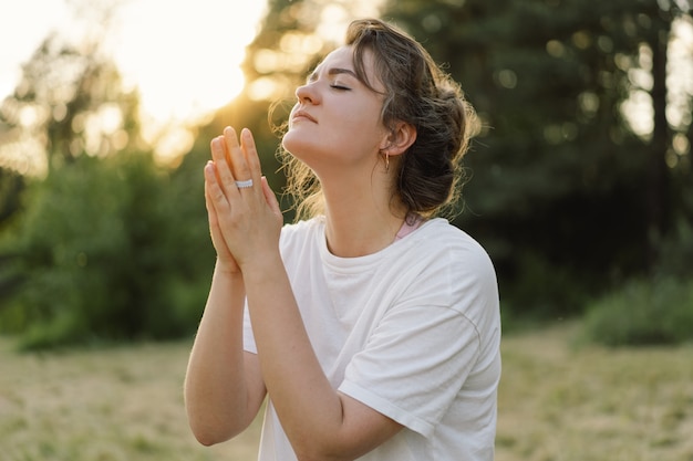 Une femme a fermé les yeux en priant dans un champ pendant un beau coucher de soleil les mains jointes dans le concept de prière pour ...