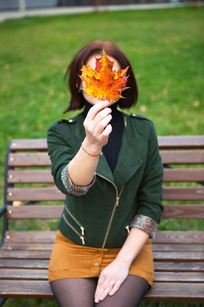 Une femme avec une feuille d'érable jaune tombée couvre son visage. Portrait d'automne sans visage sur un banc de parc. Ambiance d'automne