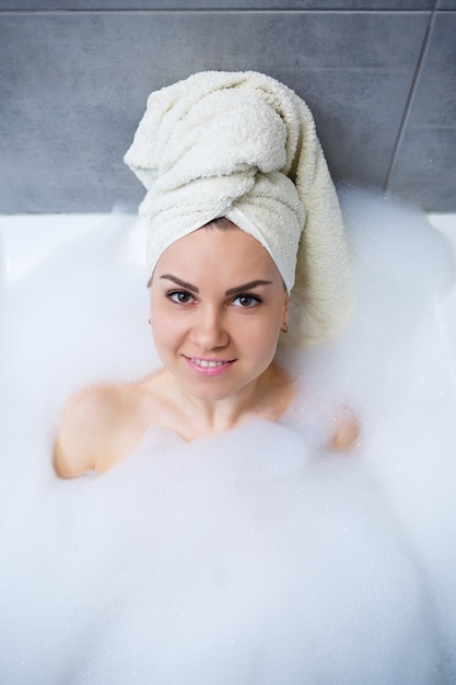Femme fille avec une serviette blanche sur la tête se trouve dans la salle de bain dans un bain blanc. Mousse pour environ. Détendez-vous après une dure journée. Soin relaxant au spa