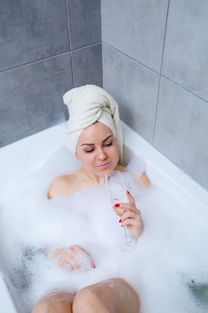 Femme fille avec une serviette blanche sur la tête se trouve avec un verre de champagne dans une baignoire blanche. Dans ses mains beaucoup de savon pipi. Mousse complète de bain à remous. Détendez-vous après une dure journée. Procédure de détente au spa