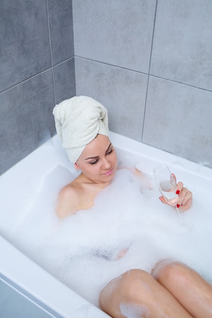 Femme fille avec une serviette blanche sur la tête se trouve avec un verre de champagne dans une baignoire blanche. Dans ses mains beaucoup de savon pipi. Mousse complète de bain à remous. Détendez-vous après une dure journée. Procédure de détente au spa