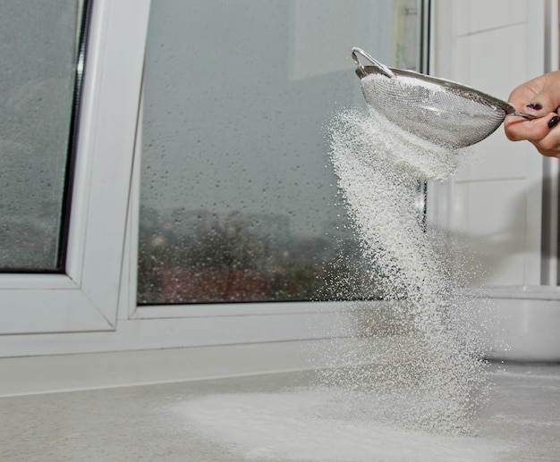 Photo femme filtrant la farine à la main à travers un tamis à la main pour la farine sur une table de cuisine blanche copier de près l'espace de cuisson de la pâte appareil de cuisine métallique