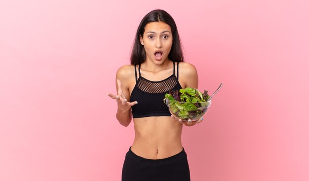 Femme fitness hispanique étonnée, choquée et étonnée par une surprise incroyable et tenant une salade