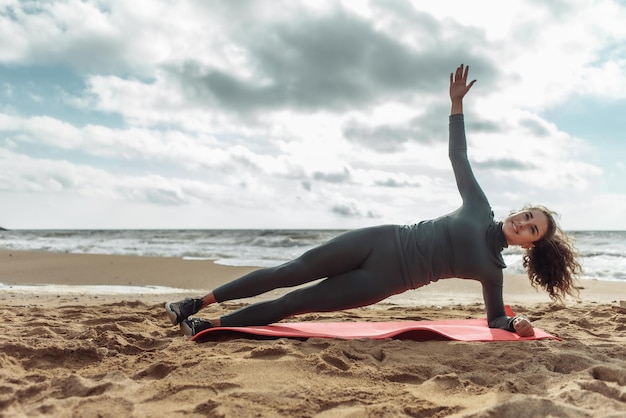 Femme fitness joyeuse aux cheveux bouclés pratiquant la planche latérale sur le sable à la plage