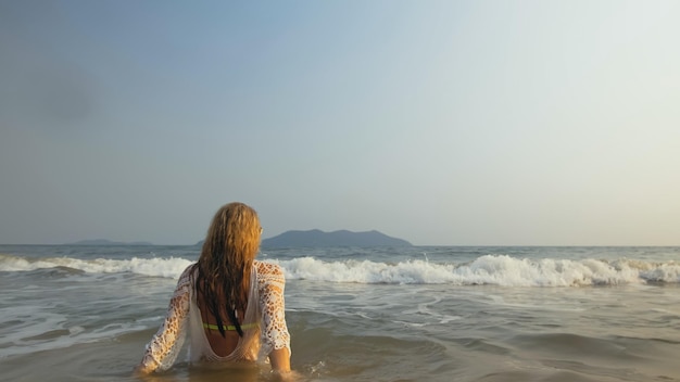 Femme folle dans une tunique blanche sur la plage près de la mer orageuse Enthousiaste blonde charmante drôle avec des cheveux bouclés mouillés dans des lunettes de soleil Souriant Happy Girl Tourist se trouve dans l'eau s'amuse Vue arrière