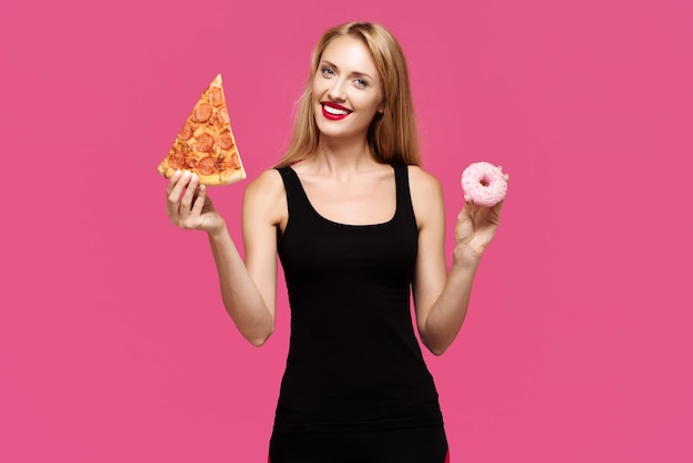 Femme sur fond rose tient une pizza et un beignet dans ses mains Le concept d'une alimentation malsaine