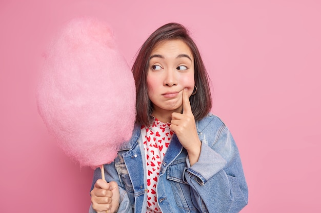 la femme force le sourire garde le doigt près du coin des lèvres regarde tristement la barbe à papa se sent seule porte une veste en jean