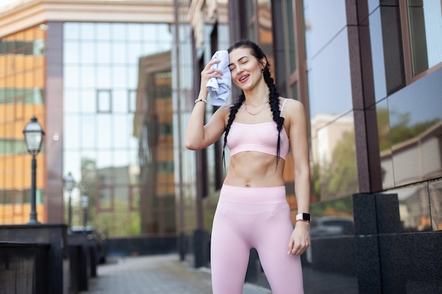 Une femme en forme fatiguée essuie la sueur avec une serviette en ville après un entraînement intense.