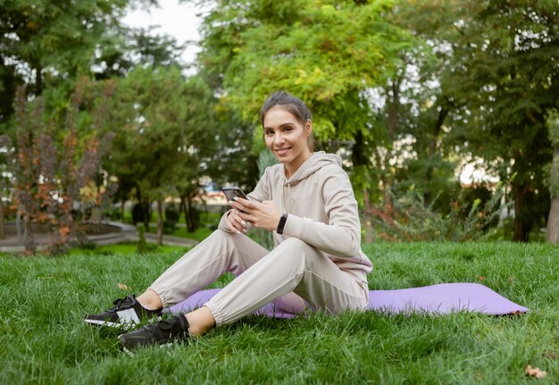 Une femme en forme heureuse a du temps de repos assise sur le tapis et utilise un smartphone dans le parc Fitness ou yoga à l'extérieur