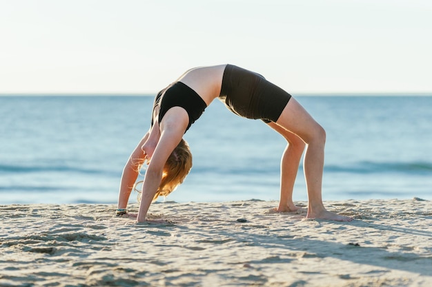 Femme en forme de race blanche faisant la position de pont du yoga sur une plage avec la mer en arrière-plan