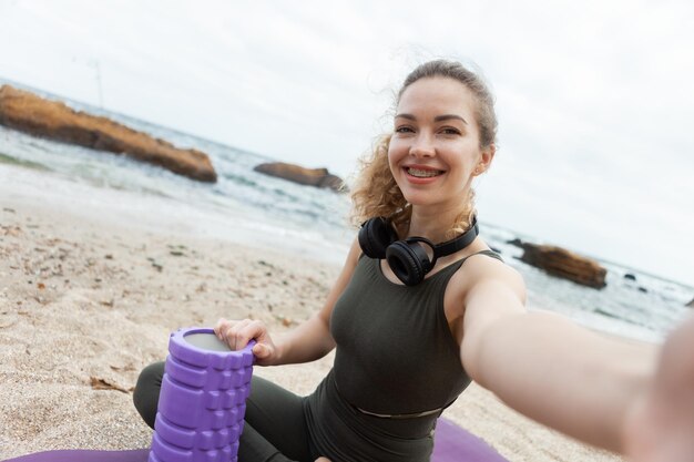 Une femme en forme avec un rouleau de massage fait du selfie tout en étant assise sur le tapis