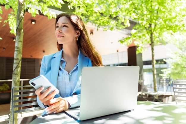 Une femme freelance attrayante assise sur son lieu de travail à l'extérieur tenant un téléphone portable à l'aide d'un ordinateur portable