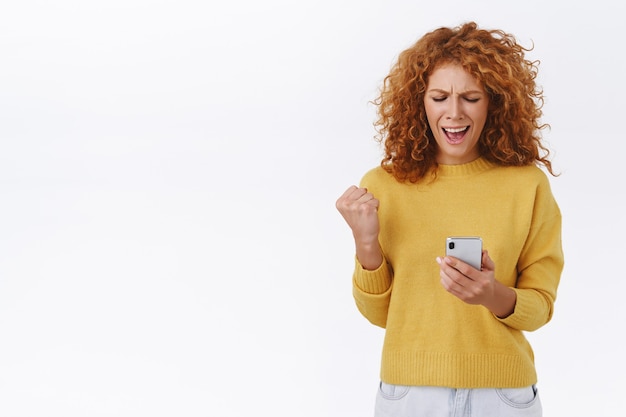 Femme frisée rousse triomphante et satisfaite, pompe à poing et disant oui, serrer le bras, tenant un smartphone, lire quelque chose de génial, obtenir des bonus dans l'application, niveau gagnant, mur blanc