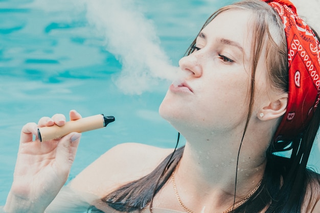 Femme fumant avec une cigarette électronique. Jeune femme fumant une cigarette électronique dans une piscine extérieure