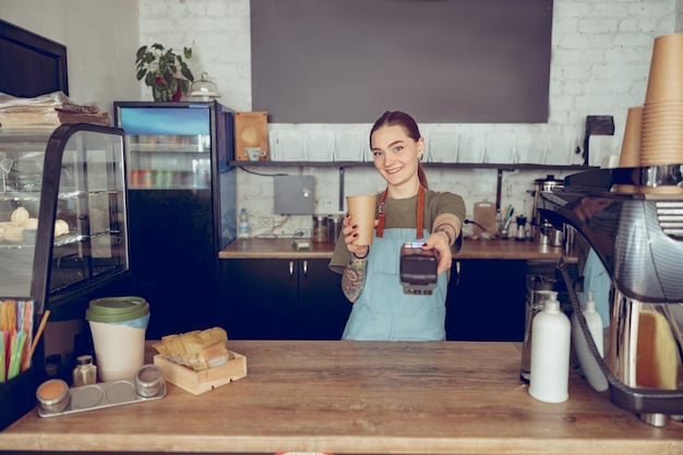 Femme gaie barista tenant un lecteur de carte sans contact et une tasse de café