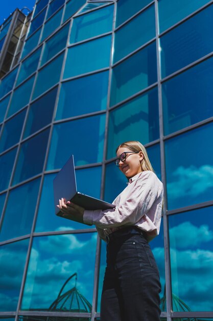 Une femme gaie concentrée dans des vêtements décontractés et des lunettes à la mode se dresse sur fond de gratte-ciel avec un ordinateur portable dans ses mains sourit et surfe sur Internet sur un ordinateur portable