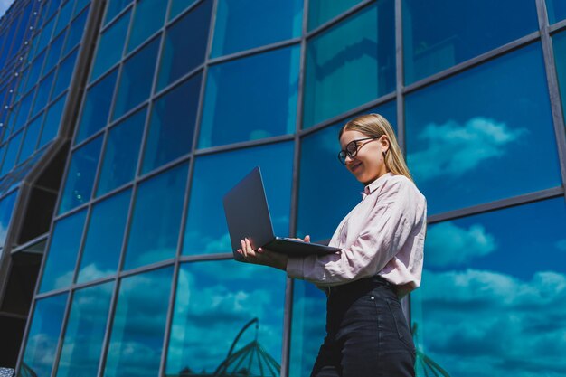 Une femme gaie concentrée dans des vêtements décontractés et des lunettes à la mode se dresse sur fond de gratte-ciel avec un ordinateur portable dans ses mains sourit et surfe sur Internet sur un ordinateur portable