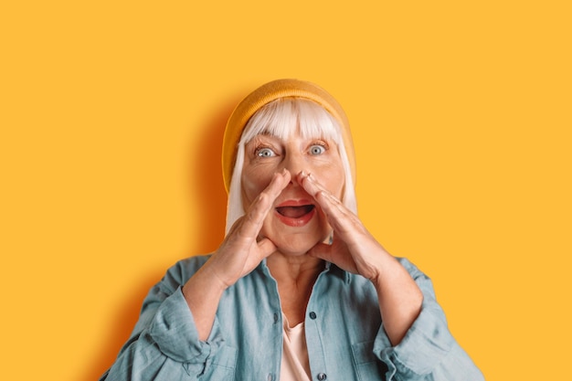 Une femme gaie dans la cinquantaine pose isolée sur un fond jaune et orange. Crier et tenir ses mains près de son visage