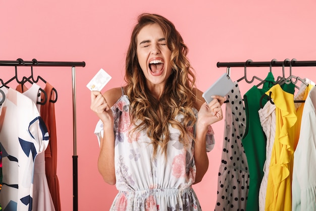 femme gaie debout près de la garde-robe tout en tenant le smartphone et la carte de crédit isolé sur rose