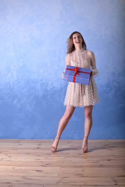 Femme gaie et heureuse dans une robe beige tenant une boîte-cadeau bleue avec un ruban rouge