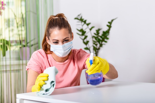 Femme avec un gant de protection et un masque facial saupoudrant de désinfectant et nettoyant la table. Restez en sécurité.