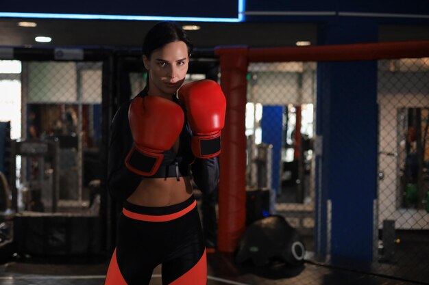 Femme en gants de boxe rouges et vêtements de sport se tient dans un rack dans la salle de boxe