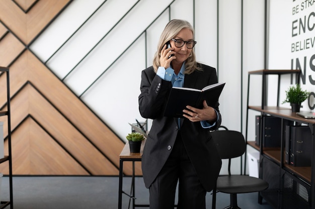 Une femme gestionnaire âgée d'un an dans un bureau stérile parle sur un téléphone portable avec un journal dans les mains