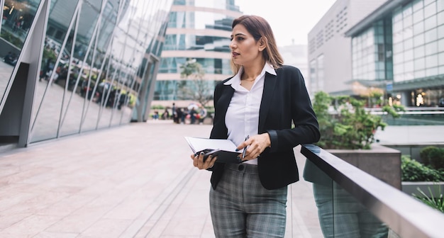 femme gestionnaire avec un pantalon gris debout près d'une clôture en verre avec un ordinateur portable