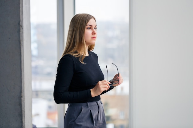 Une femme gestionnaire réussie dans son propre bureau avec de grandes fenêtres se tient debout et regarde la ville. Femme d'affaires avec des lunettes sur fond de grandes fenêtres