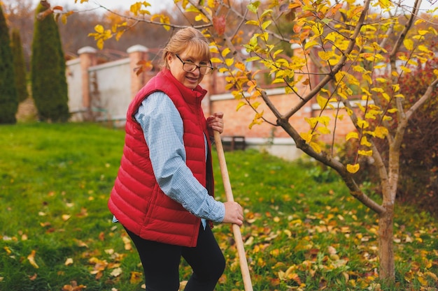 Femme en gilet rouge tenant un râteau Jardiner pendant la saison d'automne Nettoyer la pelouse des feuilles Ratisser les feuilles mortes dans le jardin