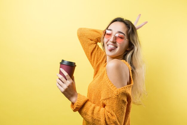 Femme glamour à lunettes dans un pull orange avec un verre de café sur fond jaune