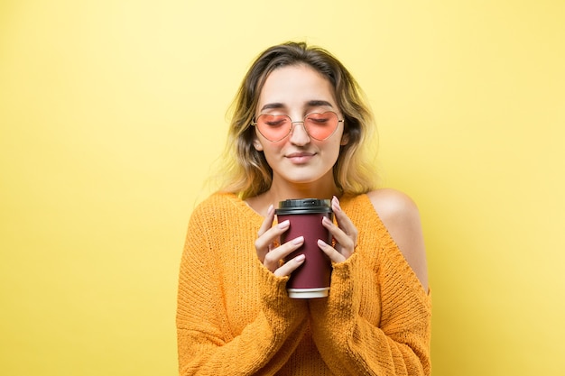 Femme glamour à lunettes dans un pull orange avec un verre de café sur fond jaune