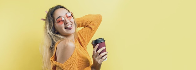 Femme glamour à lunettes dans un pull orange avec un verre de café sur fond jaune