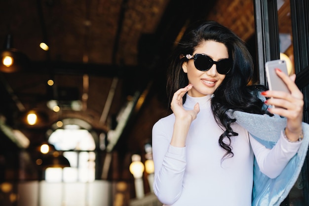 Femme glamour souriante en blouse blanche et veste à lunettes de soleil posant dans l'appareil photo de son smartphone tout en faisant un selfie en se photographiant