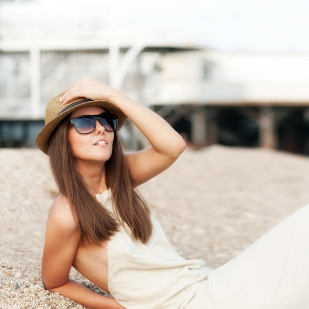 Femme habillée de mode heureuse au repos de chapeau assis sur une plage.