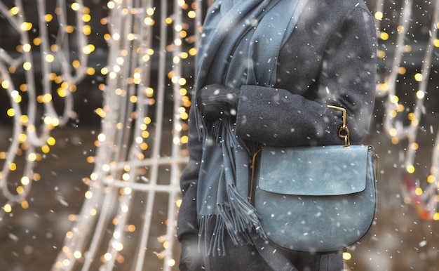 Une femme habillée à la mode porte un manteau gris et une écharpe tenant un sac à main en cuir bleu marchant pendant les chutes de neige