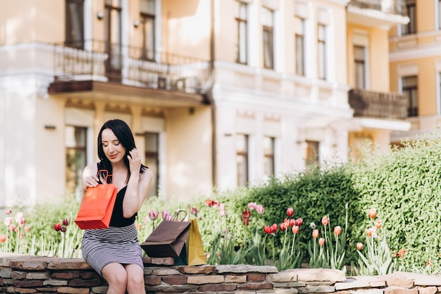 Femme habillée à la mode avec des sacs à provisions colorés assis près des fleurs dans les rues, concept d&#39;achat