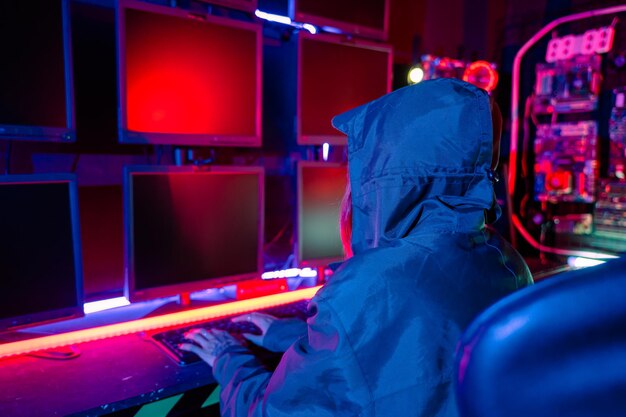 Une femme hacker porte une capuche, s'assoit devant l'ordinateur, tape sur le clavier, pirate des serveurs à la lumière du néon, joue à des jeux en ligne, salle noire, une femme hacker entre dans un centre de données.