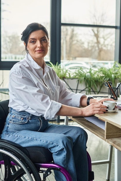 une femme handicapée de belle apparence en tenue élégante en fauteuil roulant tenant une tasse de café et regardant la caméra