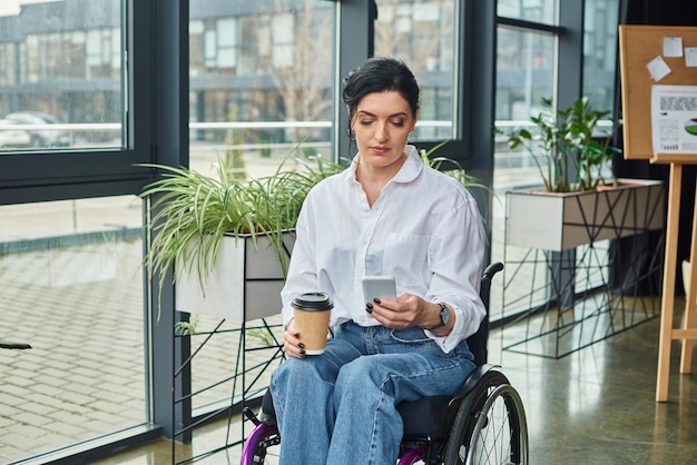 une femme handicapée brune attrayante en tenue élégante en fauteuil roulant tenant du café et un smartphone