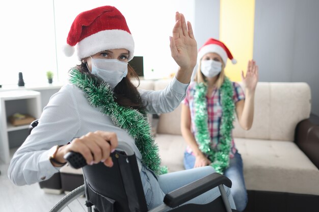 Femme handicapée en fauteuil roulant en chapeau de père Noël rouge agite sa main sur fond de son ami