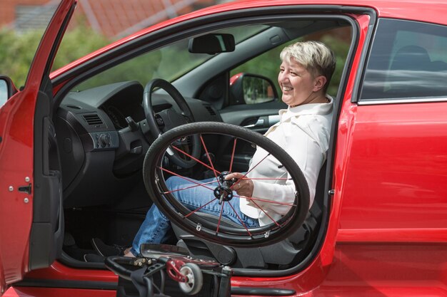 Une femme handicapée souriante assise sur le siège du conducteur et soulevant son fauteuil roulant dans la voiture