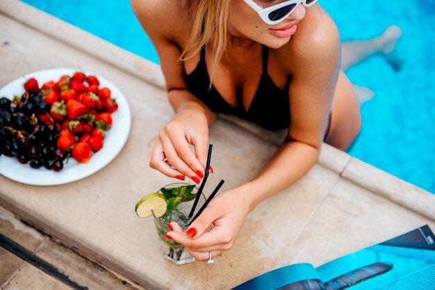 Photo une femme en haut de bikini noir boit un cocktail à côté d'une assiette de fraises.