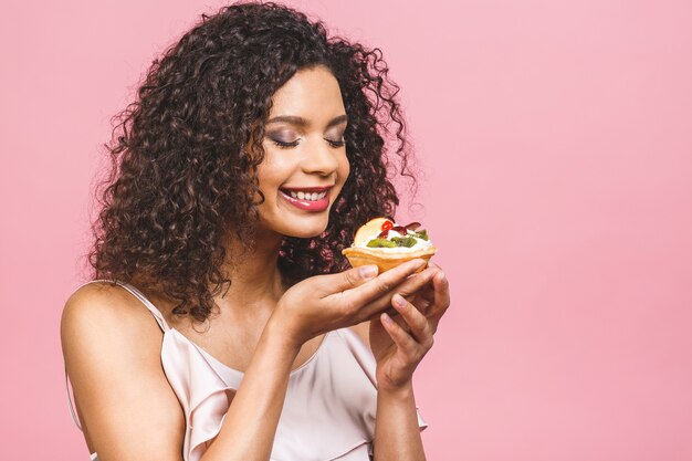 Femme heureuse afro-américaine noire avec un style de cheveux afro bouclés faisant un désordre manger un énorme dessert fantaisie sur fond rose. Manger un petit gâteau.