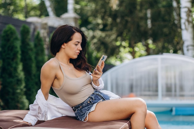 Femme heureuse à l'aide d'un téléphone intelligent au bord de la piscine de son jardin en été