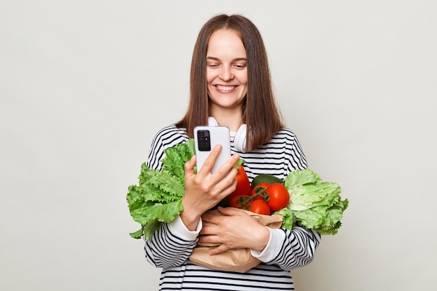Femme heureuse aux cheveux bruns portant une chemise rayée debout isolée sur fond gris commandant des légumes dans une épicerie en ligne à l'aide d'un smartphone