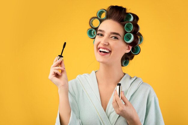 Femme heureuse avec des bigoudis tenant du mascara sur fond de studio jaune