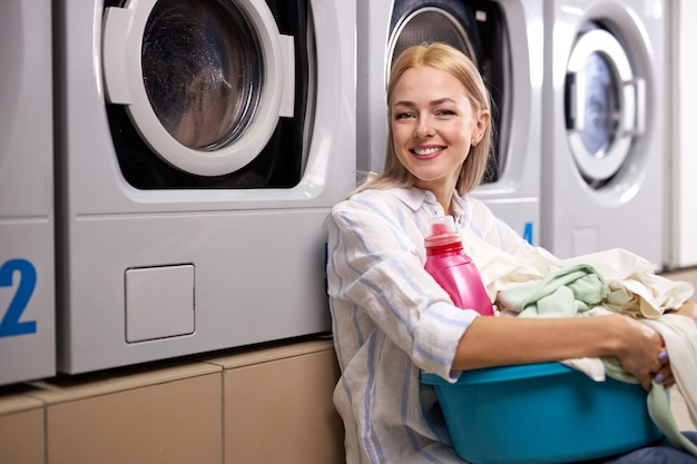 Femme heureuse blonde tenant un panier de vêtements à laver dans une buanderie automatique, la jeune femme est assise sur le sol en souriant à la caméra. lavage, nettoyage, concept de tâches ménagères
