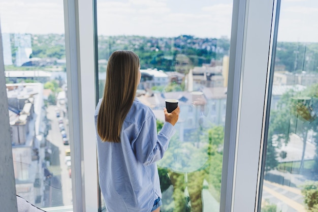 Une femme heureuse boit du café et regarde par la fenêtre dans un café Une jeune fille souriante à lunettes se tient près de la fenêtre Loisirs loisirs et temps libre Le style de vie moderne des pigistes