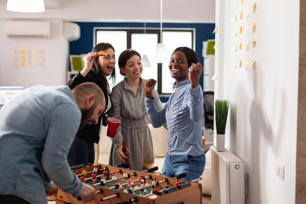 Femme Heureuse Célébrant La Victoire Du Jeu De Baby-foot à La Fête De Bureau Après Les Heures, Dégustant Des Boissons Alcoolisées Avec Des Collègues Après Le Travail. Hommes D'affaires S'amusant Avec Un Jeu D'activités De Loisirs.